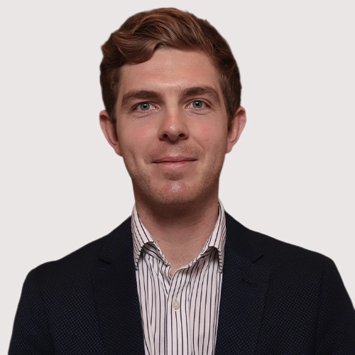 Portrait of a young man in a suit jacket and striped shirt, looking at the camera against a plain background.